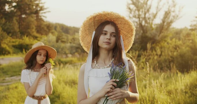 Two girl teenagers in white dresses and straw hats with flowers bouquet posing on nature background at sunny summer day