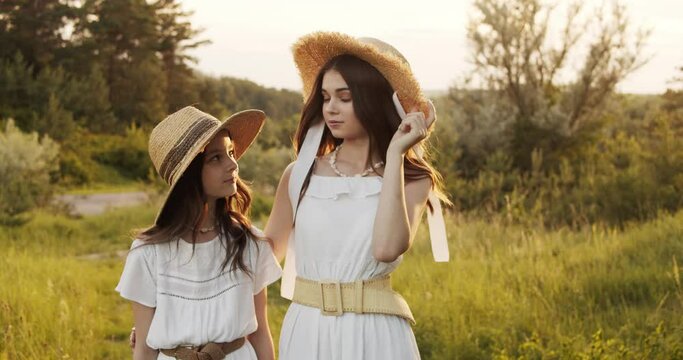 Two girl teenagers in white dresses and straw hats with flowers bouquet posing on nature landscape at sunny summer day