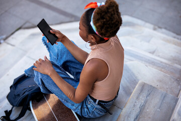 Portrait of happy african-american woman with skateboard. Young stylish woman using her phone while sitting on the stairs