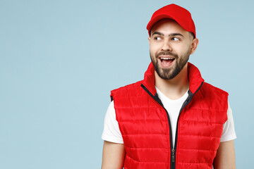 Professional delivery guy employee man in red cap white T-shirt vest uniform workwear work as dealer courier look aside on promo area isolated on pastel blue color background studio. Service concept.