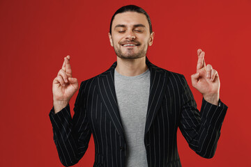 Young businessman latin man 20s in black striped jacket grey shirt waiting for special moment, keeping fingers crossed, making wish, eyes closed isolated on red background Concept of people lifestyle