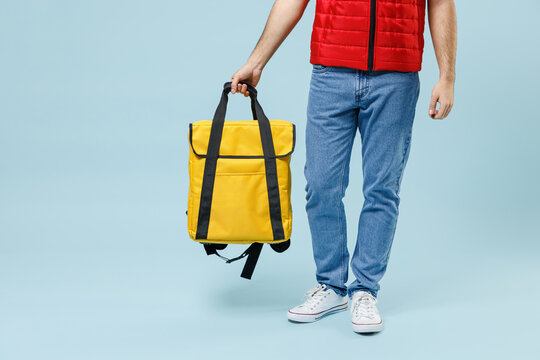 Delivery guy employee man in red vest uniform jeans on legs workwear work dealer courier hold in arm yellow thermal food bag backpack isolated on pastel blue color background studio Service concept
