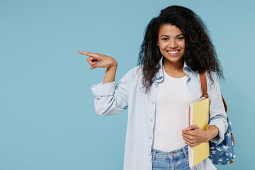 Young fun african american girl teen student in denim clothes backpack hold books point finger aside copy space mock up isolated on blue background Education in high school university college concept - obrazy, fototapety, plakaty