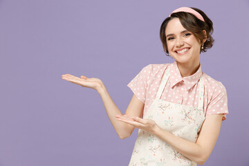 Young happy smiling housewife housekeeper chef cook baker woman wear pink apron point hand aside on workspace area mock up isolated on pastel violet background studio portrait. Cooking food concept