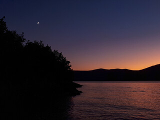 Fototapeta na wymiar Moon and tree silhouettes at the sea against evening sky on Krk, Croatia. Night at the beach