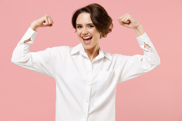 Young strong fitness successful employee business woman corporate lawyer in classic formal white shirt work in office show hand biceps muscles demonstrating strength isolated on pastel pink background