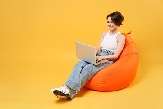 Young smiling woman 20s with bob haircut wearing white tank top shirt using laptop pc computer chat online browsing surfing internet sit in orange bag chair hold face isolated on yellow background