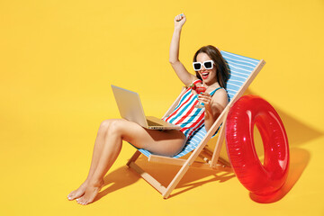 Full body length happy young woman wear red blue one-piece swimsuit sit on chair work hold laptop pc isolated over vivid yellow color wall background studio Summer hotel pool sea rest sun tan concept