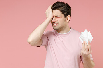 Unhealthy sick allergic crying man has watery swelling red eyes runny nose suffer from allergy symptoms hay fever hold paper napkin isolated on pastel pink color background studio. Seasonal rhinitis.