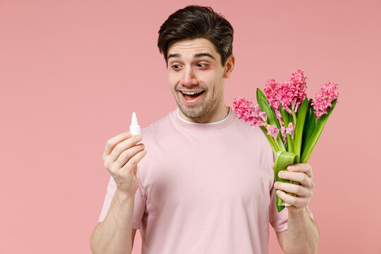 Sick unhealthy ill allergic man has red eyes runny stuffy sore nose suffer from allergy symptoms hay fever hold blooming flower plant uses nasal drops isolated on pastel pink color background studio.
