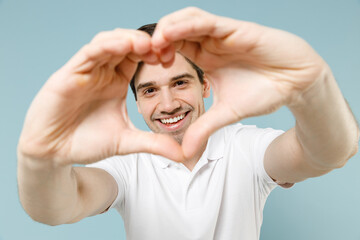 Young lovely caucasian man 20s in white casual basic t-shirt showing shape heart with hands look through heart-shape sign isolated on pastel blue background studio portrait People lifestyle concept.