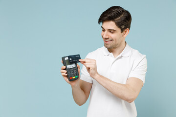 Young smiling happy man wearing white casual basic t-shirt holding wireless modern bank payment terminal to process and acquire credit card payments isolated on pastel blue background studio portrait. - obrazy, fototapety, plakaty