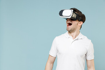 Young amazed surprised shocked man 20s in white casual basic t-shirt play pc game watching in vr headset pc gadget isolated on pastel blue color background studio portrait People lifestyle concept.
