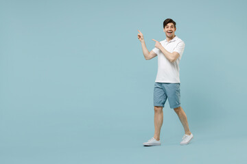 Full length young fun surprised man wear white basic t-shirt point finger aside on workspace area copy space mock up isolated on pastel blue color background studio portrait. People lifestyle concept
