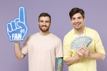 Two young excited happy successful men friends together in casual t-shirt fan foam glove finger holding fan of cash money in dollar banknotes isolated on purple background. People lifestyle concept