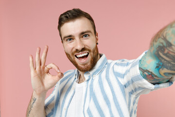 Fototapeta premium Close up young happy caucasian unshaven man 20s in blue striped shirt doing selfie shot on mobile cell phone show ok okay gesture isolated on pastel pink background studio. People lifestyle concept.