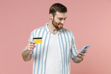 Young caucasian unshaven man in blue striped shirt use mobile cell phone hold credit bank card do...