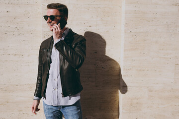 Young joyful fun trendy man in leather jacket shirt glasses stand on outdoors building wall bakcground, talk by mobile cell phone spread hands look aside Concept of people urban lifestyle and clothes