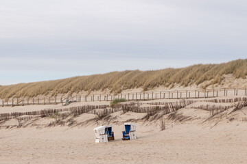 empty beach chairs stand on an empty beach directly behind a dune and a wooden jetty. 
