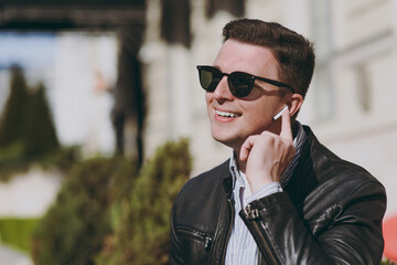 Young smiling man in black leather jacket shirt glasses stand outdoors, talk by mobile cell phone use air pods headphones, listen to music walking in city downtown Concept of people urban lifestyle