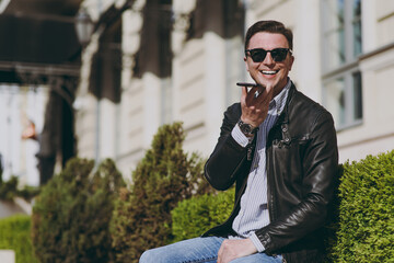 Young stylish happy fun smiling man in black leather jacket shirt glasses stand outdoors, talk by mobile cell phone record voice message walking in city downtown. Concept of people urban lifestyle