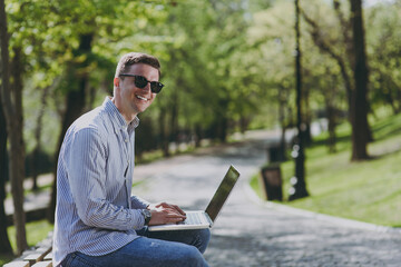 Fototapeta premium Side view young freelancer smiling happy student man in shirt glasses sitting on bench in green park work by laptop pc computer outdoors distance learning and remote job. Concept of urban lifestyle