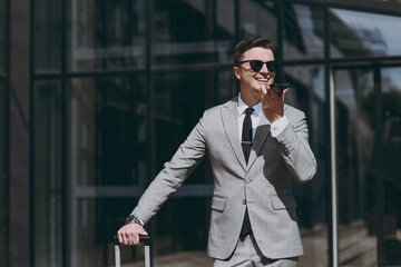 Young employee business man in grey suit stand near office glass wall building outdoors in downtown city talk use mobile cell phone send voice message hold valise, booking taxi, Business trip concept