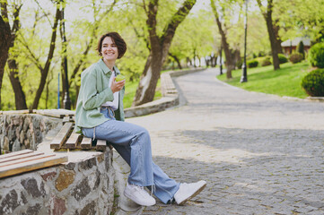 Fototapeta na wymiar Young smiling happy student woman 20s in casual green jacket jeans sitting on bench in city spring park outdoors resting eat apple fruit look aside. People vegeterian healthy urban lifestyle concept
