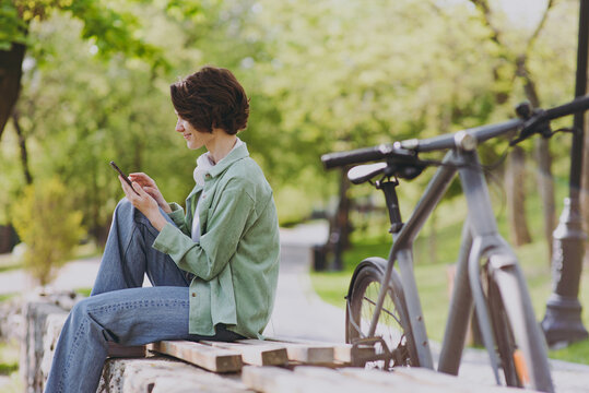 SIde View Happy Young Woman 20s In Green Jacket Jeans Sit On Bench Near Bicycle In City Spring Park Outdoors Hold Mobile Cell Phone Chat Online In Social Network People Active Urban Youthful Concept