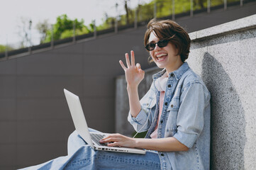 Side view freelancer young fun student woman in jeans clothes headphones eyeglasses listen to music leaning on building wall sit on concrete steps outdoors use laptop pc computer show ok okay gesture.