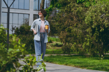Full body length young happy woman in jeans clothes strolling near building walk in park outdoors hold takeaway paper cup coffee to go, laptop pc computer look aside People urban lifestyle concept