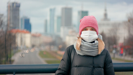 COVID-19 Coronavirus Pandemic or Smog Background. Portrait of Young Woman in Surgical Face Mask Staying Outdoors in Big City and Looking to Camera. Picture with Copy Space