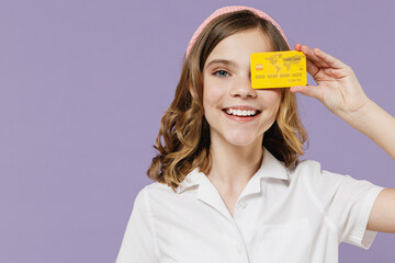 Little cheerful kid girl 12-13 years old in white shirt holding credit bank card cover eye with credit bank card isolated on purple background children studio portrait. Childhood lifestyle concept