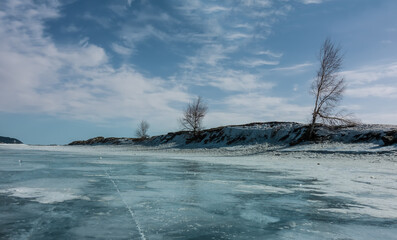Several trees grow on the shore of the frozen lake. Bare trunks and branches against a blue sky with cirrus clouds. There is snow on the ice, cracks. Baikal