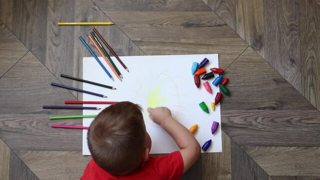 little boy in red t-shirt drawing the sun with pencils or crayons lying on the floor in the living room, top view. children education development concept