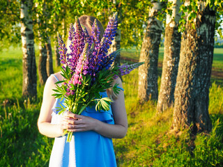Beautiful girl holding and hiding her face behind bouquet of summer flowers of purple lupins. Young happy woman enjoying nature. Hello summer. Wellness, harmony and freedom