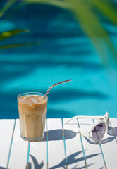 Ice coffee Cyprus Frappe Fredo against blue clear water of the swimming pool, on white table, with sunglasses. Summer minimalistic background, holiday or vacation concept. Sun and shadows.Copy space