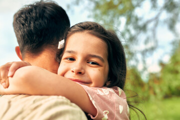 Rear view of a father hugging his smiling daughter looking to the camera spending time together in the park. Cute little girl embracing her father.