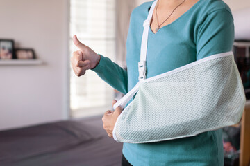Patient woman showing thump up while wearing arm sling for relieve broken arms at home