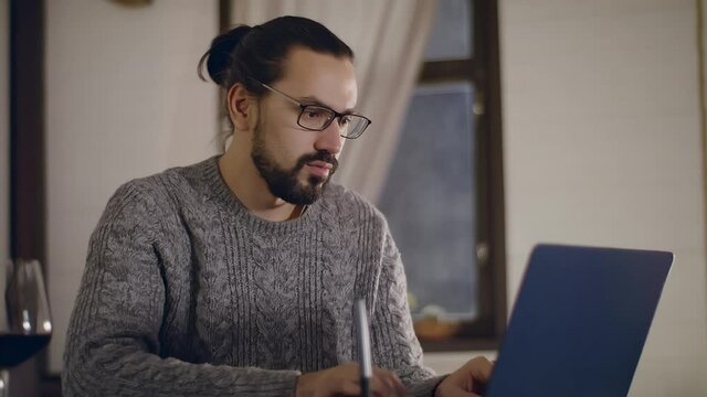 A young, handsome man in glasses works on a laptop with a glass of wine.