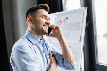 Smiling businessman in formal wear talking on mobile phone near blurred flipchart