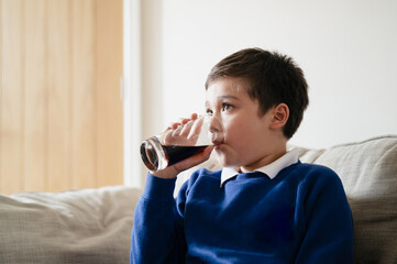 Portrait schoolboy drinking soda or soft drink with glass,Child enjoying cold fizzy dink while watching TV after back from school,Kid relaxing at home in hot sunny day summer. Unhealthy food concept