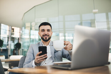 Cheerful businessman with smartphone and laptop having cup of coffee
