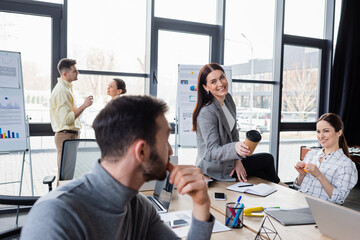 Obraz premium Cheerful businesswoman holding paper cup near blurred colleague during meeting