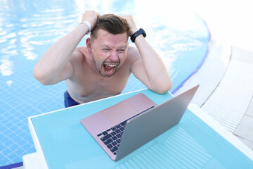 Man screaming and holding his hair and looking at laptop screen on side of pool