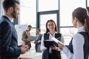 Smiling businesswoman holding notebook near colleagues with gadgets in office