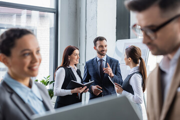 Cheerful businessman pointing with finger near colleagues with notebook and digital tablet in office