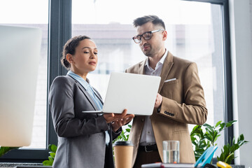 African american businesswoman holding laptop and talking with colleague near blurred coffee and water
