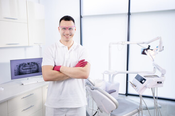 Portrait of a young smiling dentist in work glasses and in a white uniform. The man folded his arms and stands at the dental chair in the office