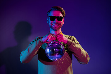 Photo portrait young man in sunglass smiling keeping disco ball isolated gradient violet color...